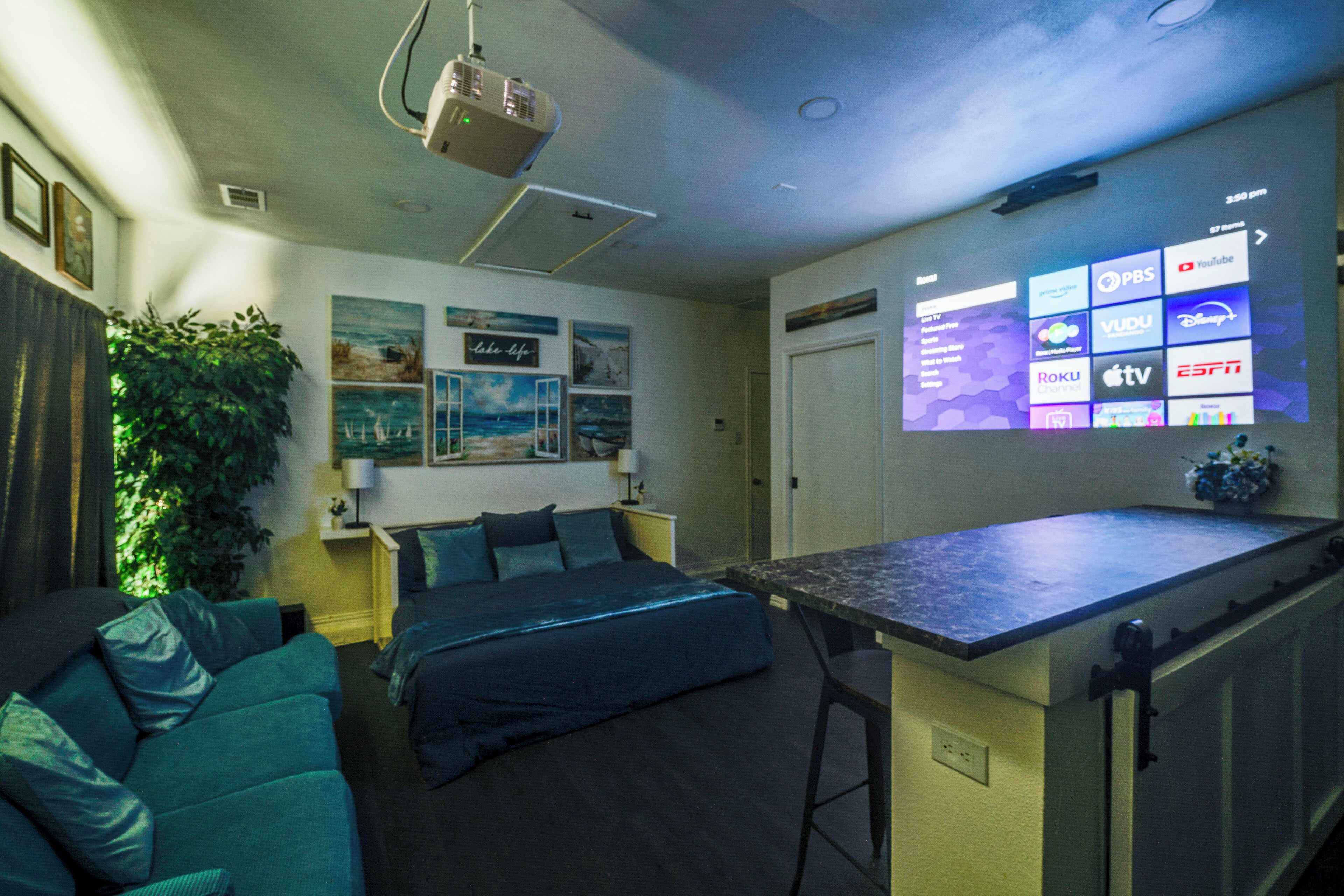 A modern hotel room with a bed, a sofa, a small table, and a TV displaying channel options.