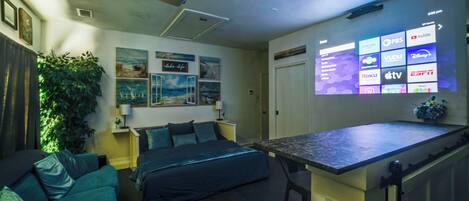 A modern hotel room with a bed, a sofa, a small table, and a TV displaying channel options.