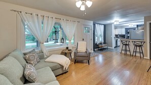 A spacious living area with a grey sofa, a white armchair, and a wooden floor. A kitchen with a bar area is visible in the background. - Dry Ridge Rental Home w/ Hot Tub & Game Room! (Crittenden)