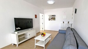 A modern living room with a flat-screen TV on a white entertainment unit, a grey sofa, a small white coffee table with fruit, and a white cabinet with drawers. - Ferienwohnung Ocean Sounds A in Gran Tarajal, Innenstadt, Meerblick, Terrasse (Gran Tarajal)