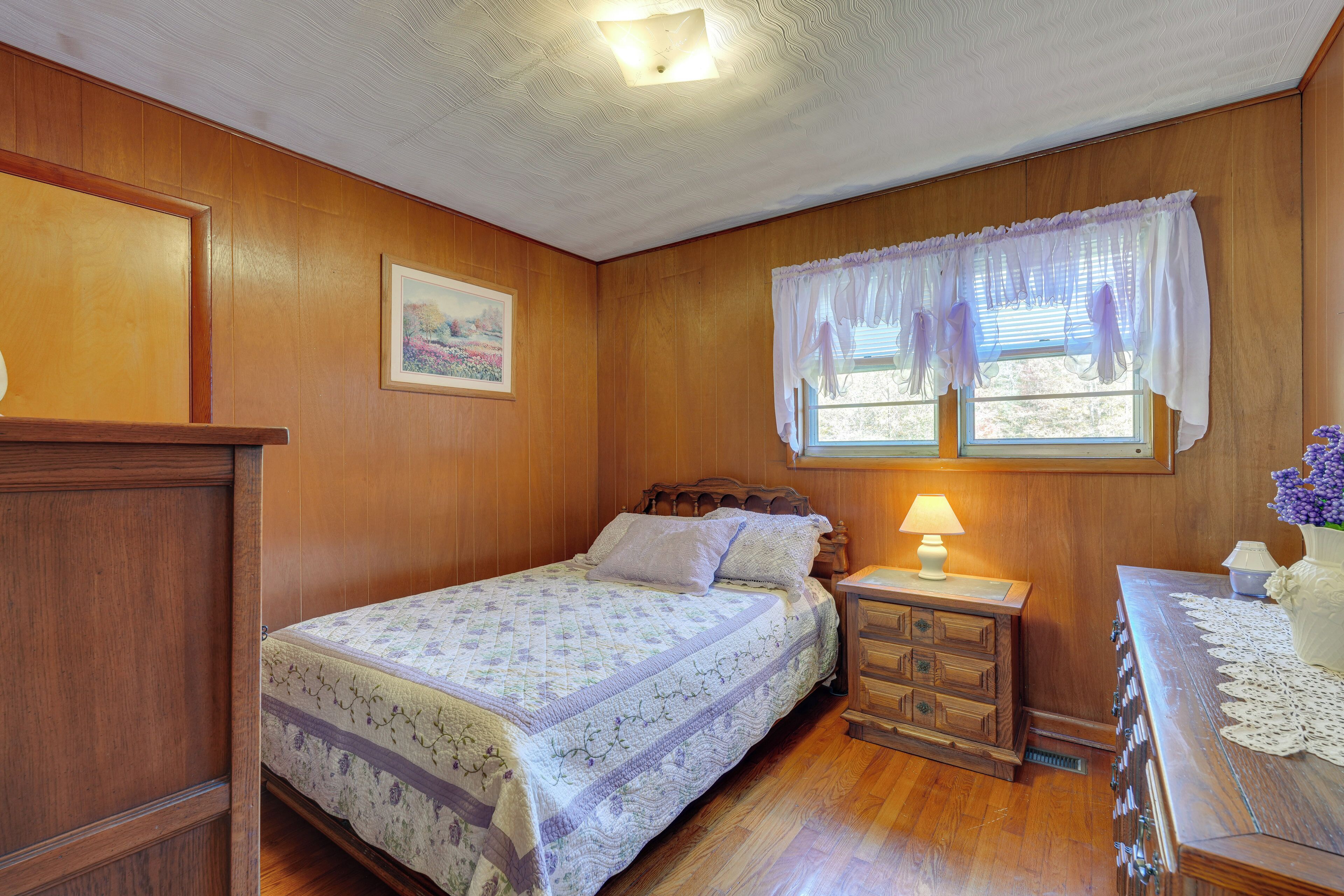 A bedroom with a wooden paneled wall, a bed with a floral quilt, a nightstand with a lamp, and a dresser with a vase of flowers.
