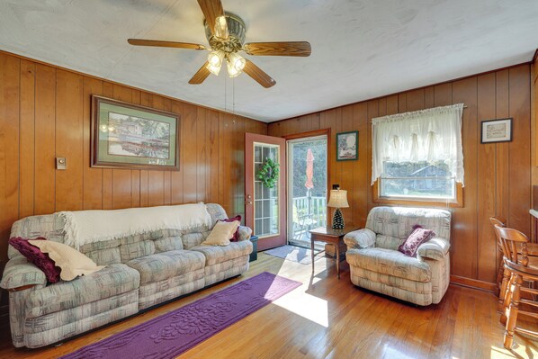 A cozy living room with wooden walls, a ceiling fan, a sofa, a chair, and a dining table.