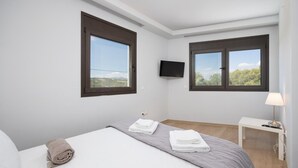 Een hotelkamer met twee grote ramen, een bed met wit en grijs beddengoed, een flatscreen televisie aan de muur en een nachtkastje met een lamp.
