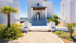 Un edificio moderno blanco con una escalera central que lleva a una puerta, rodeado de vegetación y un cielo azul claro.