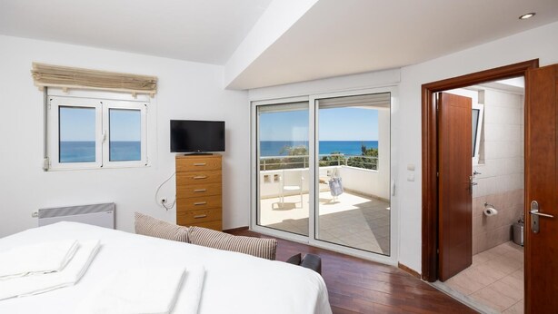 A bedroom with a bed, a TV on a wooden cabinet, a balcony with a view of the sea, and a sliding door to a bathroom.
