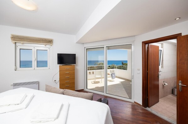 A bedroom with a bed, a TV on a wooden cabinet, a balcony with a view of the sea, and a sliding door to a bathroom. - Villa Pelagos Blue - Four Bedroom Villa, Sleeps 8 (Rhodes)