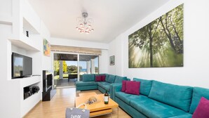 A modern living room with a teal sofa, a wooden coffee table, a TV, and a painting of a forest.
