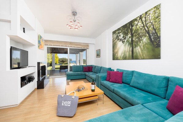 Ein modernes Wohnzimmer mit einem petrolfarbenen Sofa, einem Holztisch, einem Fernseher und einem Waldgemälde.