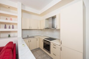 A modern kitchen with beige cabinets, a stainless steel range hood, a double sink, and a built-in oven. There is a red chair and a small green vase on the counter.