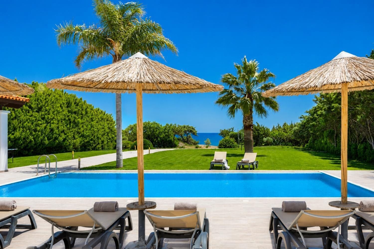 Una piscina con sillas de descanso y sombrillas de paja, rodeada de vegetación y una vista despejada al mar.
