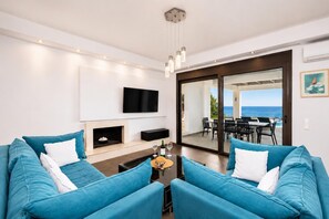 A modern living room with a sofa set, a flat-screen TV, a fireplace, and a dining area with a view of the ocean.