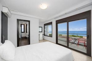 A hotel room with a bed, a desk, a chair, a balcony with a view of the sea, and a sliding glass door.