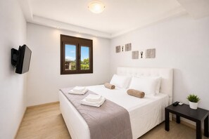 A modern bedroom with a bed, a TV mounted on the wall, a window with a view, and a small table with a plant.