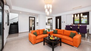 Een moderne woonkamer met een hoekbank, een bijzettafeltje met fruit en drankjes, een kroonluchter en een eethoek met paarse stoelen.