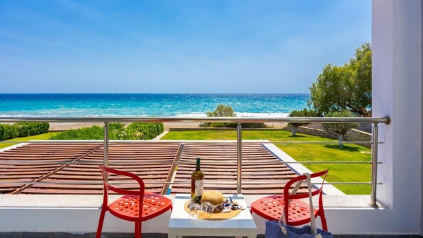 Een balkon met uitzicht op de oceaan, twee rode stoelen, een tafel met een fles en glazen, en een hoed.