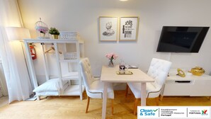 Ein sauberes und modernes Zimmer mit einem weißen Bett, einem kleinen Tisch mit Stühlen, einem Fernseher und gerahmten Bildern an der Wand.