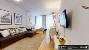 Ein modernes Wohnzimmer mit einer braunen Couch, einem weißen Sessel und einem Bett mit weißem Kopfteil. An der Wand ist ein Fernseher angebracht, und darüber hängt ein dekorativer Spiegel.