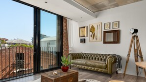 A spacious living room with a leather sofa, a wooden coffee table with a plant, a large window with a view of a brick building, and a tripod lamp. - The Signature 102 (Cape Town)