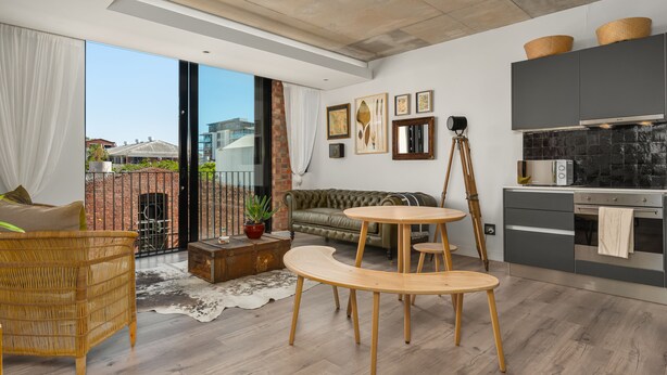 A modern living room with a large sliding glass door, a sofa, a wooden dining table, and a kitchen area with dark cabinets and a stove. - The Signature 102 (Cape Town)