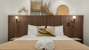 A neatly made bed with a white duvet, a beige throw blanket, and two rolled white towels. The headboard is wooden with vertical slats and two wall-mounted light fixtures. - The Signature 102 (Cape Town)