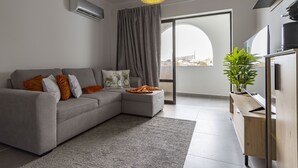 A modern living room with a grey sofa, a wooden side table, and a large window with a view of the city.