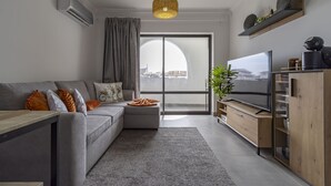 A modern living room with a grey sofa, a wooden TV stand, and a large window with a city view.