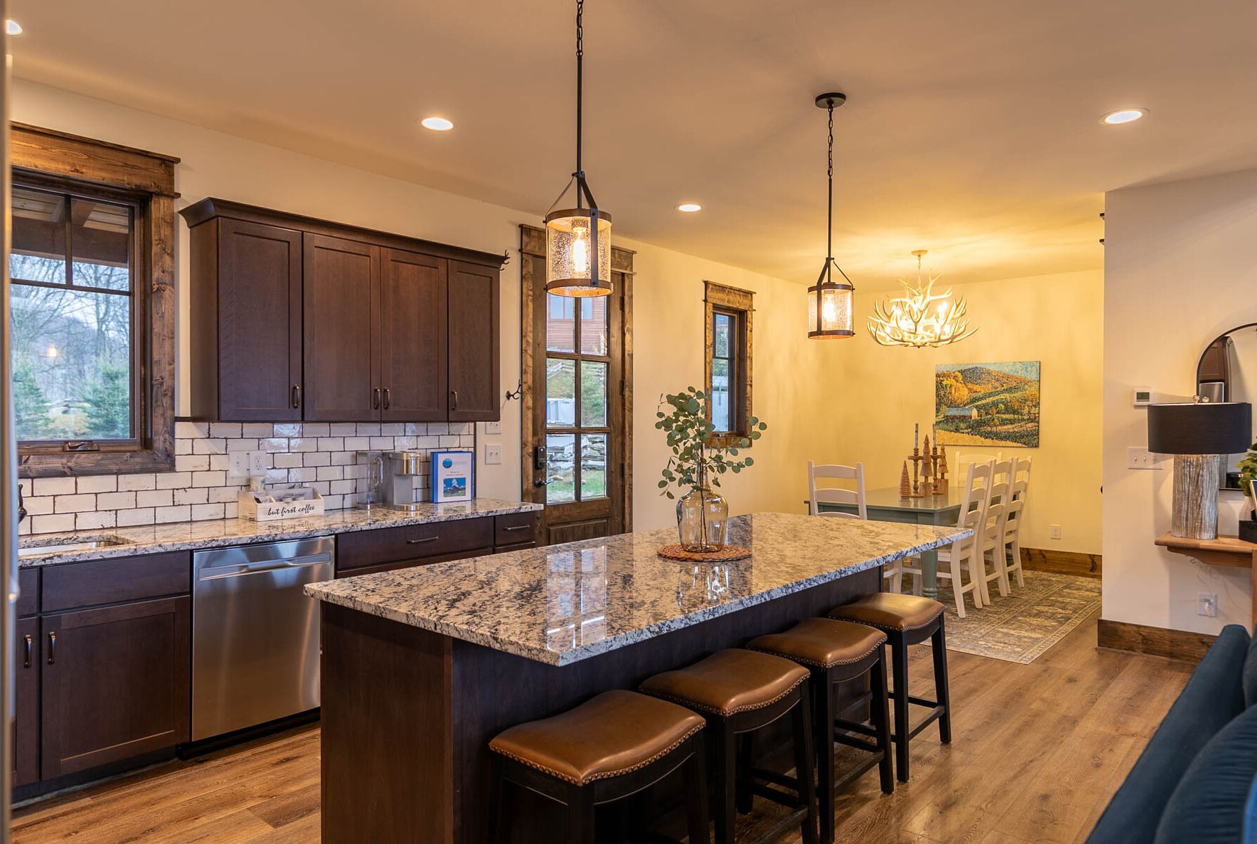 A modern kitchen with dark wood cabinets, a granite island with bar stools, pendant lighting, and a dining area with a chandelier.