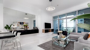 A modern living room with a glass coffee table, a grey sofa, and a large window offering a city view.