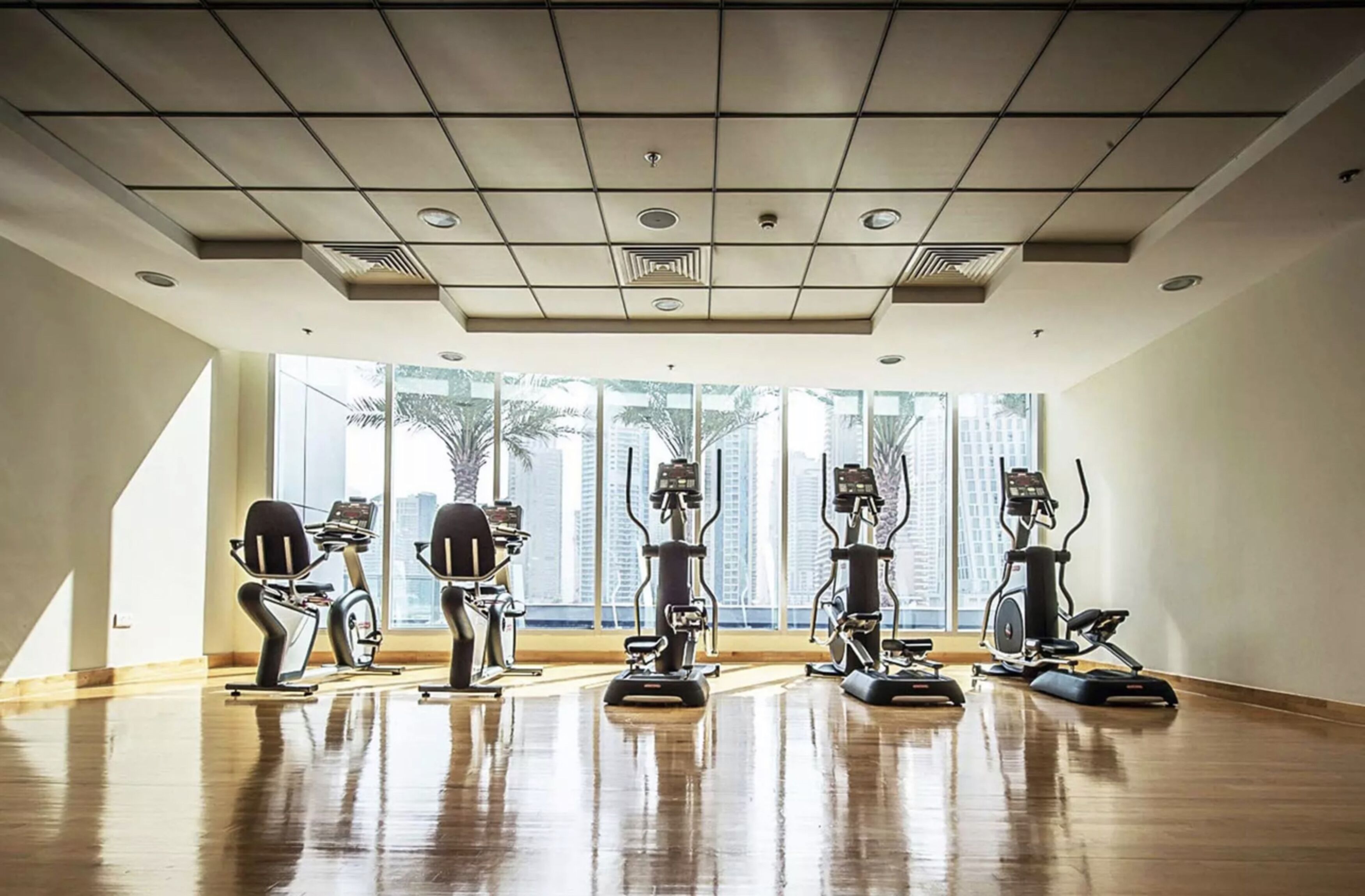 A gym with exercise bikes and treadmills arranged in a row, large windows with a city view, and a wooden floor.