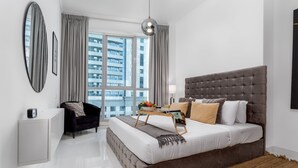 A modern bedroom with a large bed, a side table, a chair, and a view of the city through large windows.
