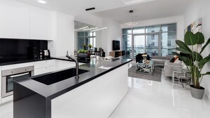 A modern kitchen with a central island, a dining area with a glass table, and a living room with a sofa and a large plant.