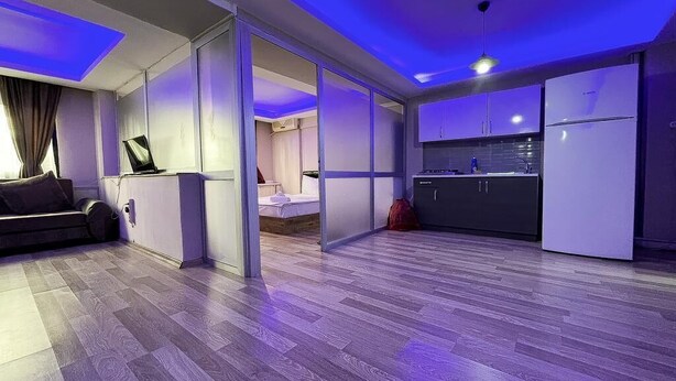 A modern living room with a sofa, a coffee table, and a laptop on a desk. There is a kitchen area with cabinets and a refrigerator. - De La Elpida Otel (Izmir)