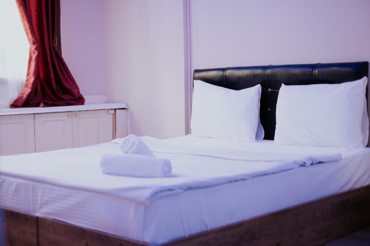 A neatly made bed with white linens, a black headboard, and a red curtain on the window.