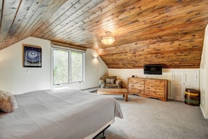 A bedroom with a sloped ceiling, a bed, a dresser, a television, and a window with a view of trees. - 1107 American Way (Breckenridge)
