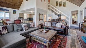 A spacious living room with a large couch, a coffee table, and a leather sofa. There is a staircase leading to an upper level, and a kitchen is visible in the background. - 1107 American Way (Breckenridge)