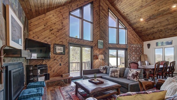 A spacious living room with a high ceiling, wooden walls, and large windows. There is a fireplace, a television, and a dining area with chairs and a table. - 1107 American Way (Breckenridge)