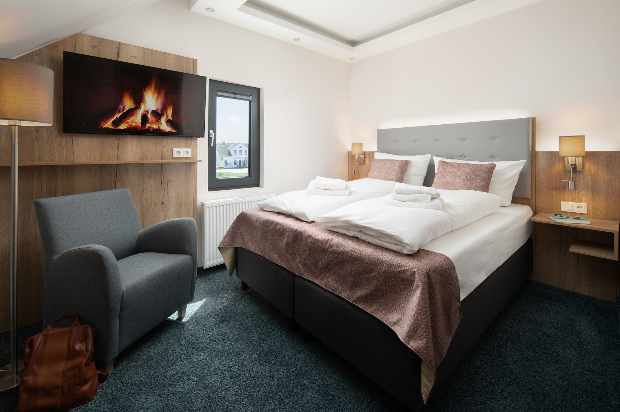 A modern hotel room with a large bed, a comfortable armchair, a flat-screen TV mounted on the wall, and a window with a view of a building.
