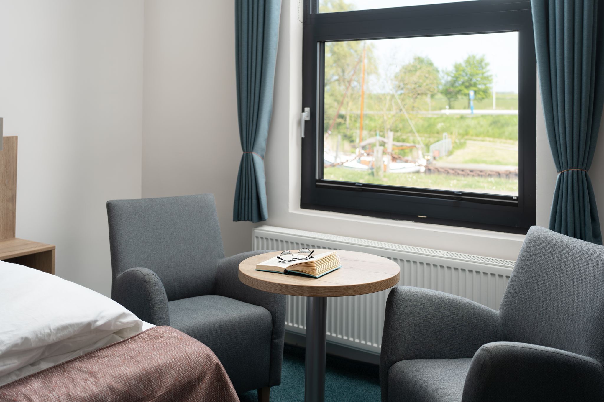 A hotel room with a bed, two grey armchairs, a small round table with a book and glasses, and a window with curtains.