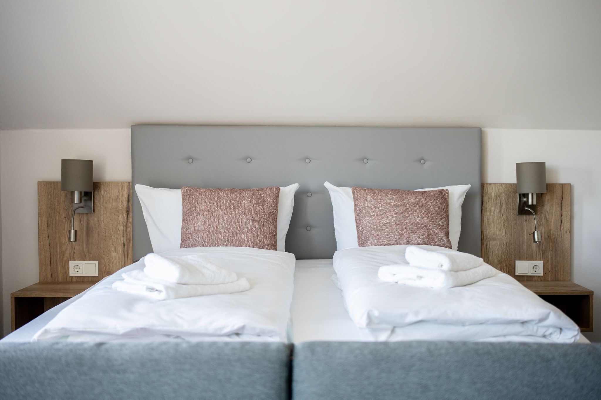 A double bed with white linens and two pillows, a grey headboard, and wooden nightstands with lamps.