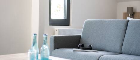 En moderne stue med en grå sofa, et lille træbord med to glasflasker, og et vindue med persienner.