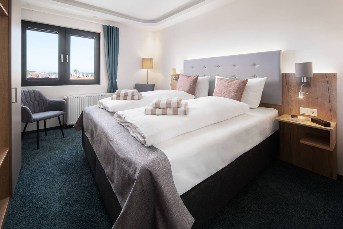 Ein Hotelzimmer mit zwei Betten, Stadtblick, einem Sessel und einem Nachttisch mit Lampe.
