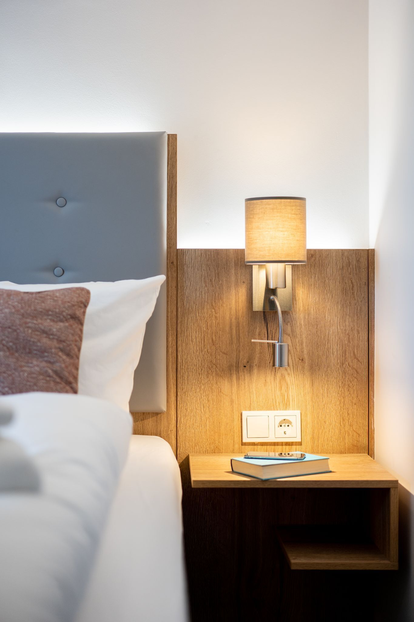 Una cama bien hecha con un cabecero gris, una mesita de noche de madera con una lámpara, y un libro sobre la mesa.