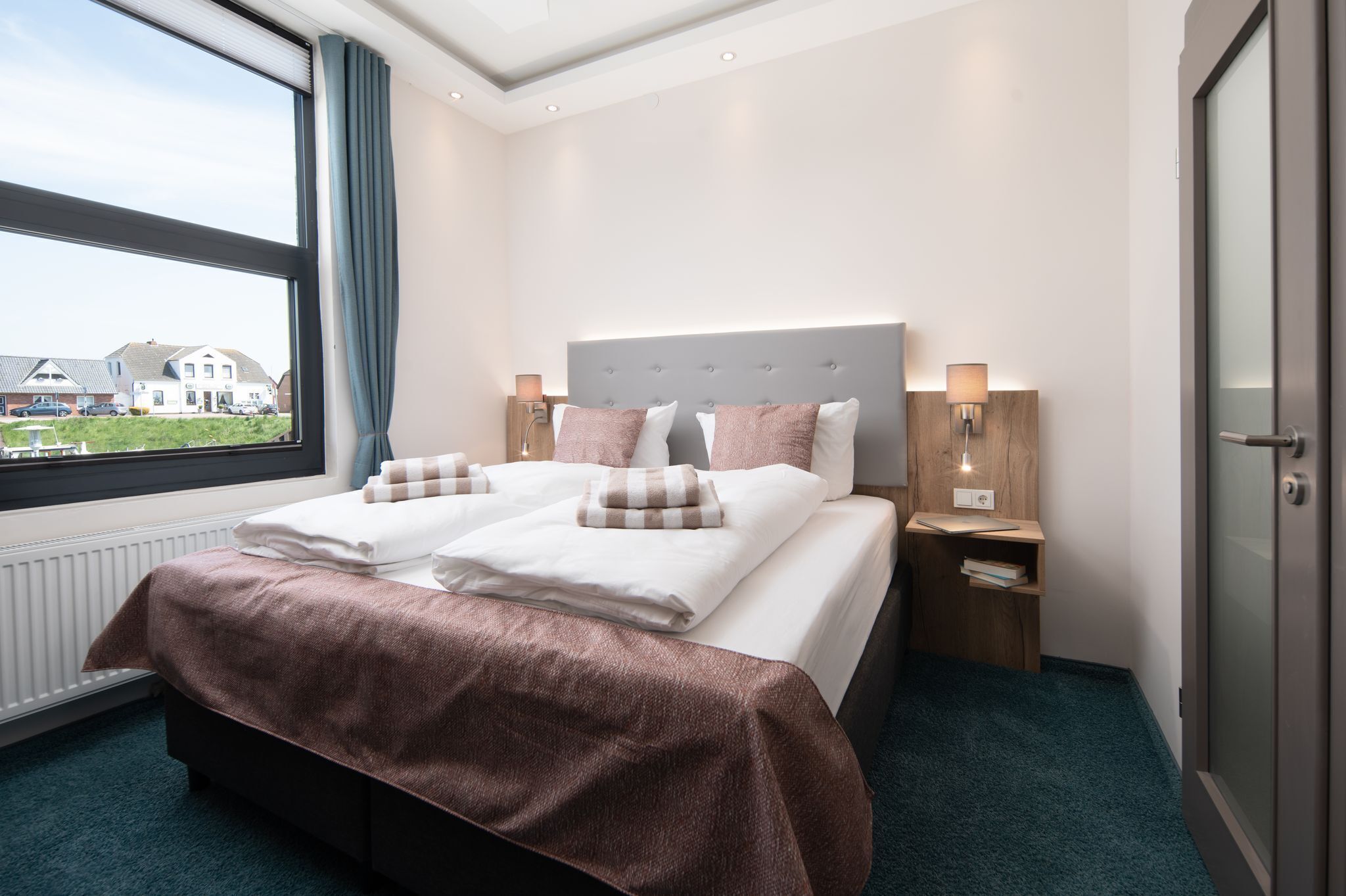 Habitación de hotel con una cama grande, dos mesitas de noche, una ventana con cortinas y vistas a casas.