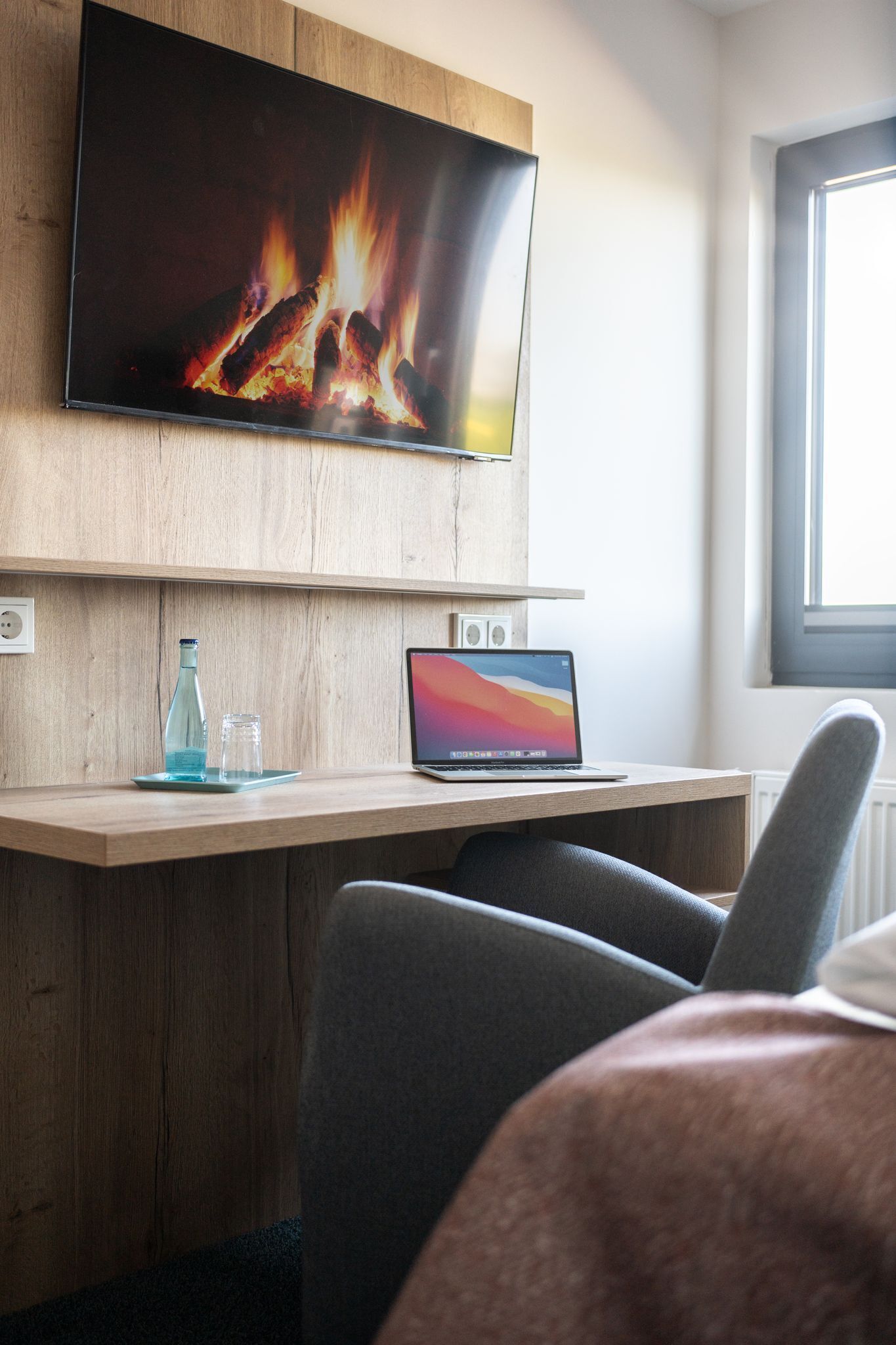 A modern room with a flat-screen TV mounted on a wooden wall, a laptop on a wooden desk, and a window providing natural light.