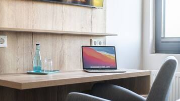 Een moderne kamer met een flatscreen televisie aan een houten wand, een laptop op een houten bureau, en een raam dat voor natuurlijk licht zorgt.