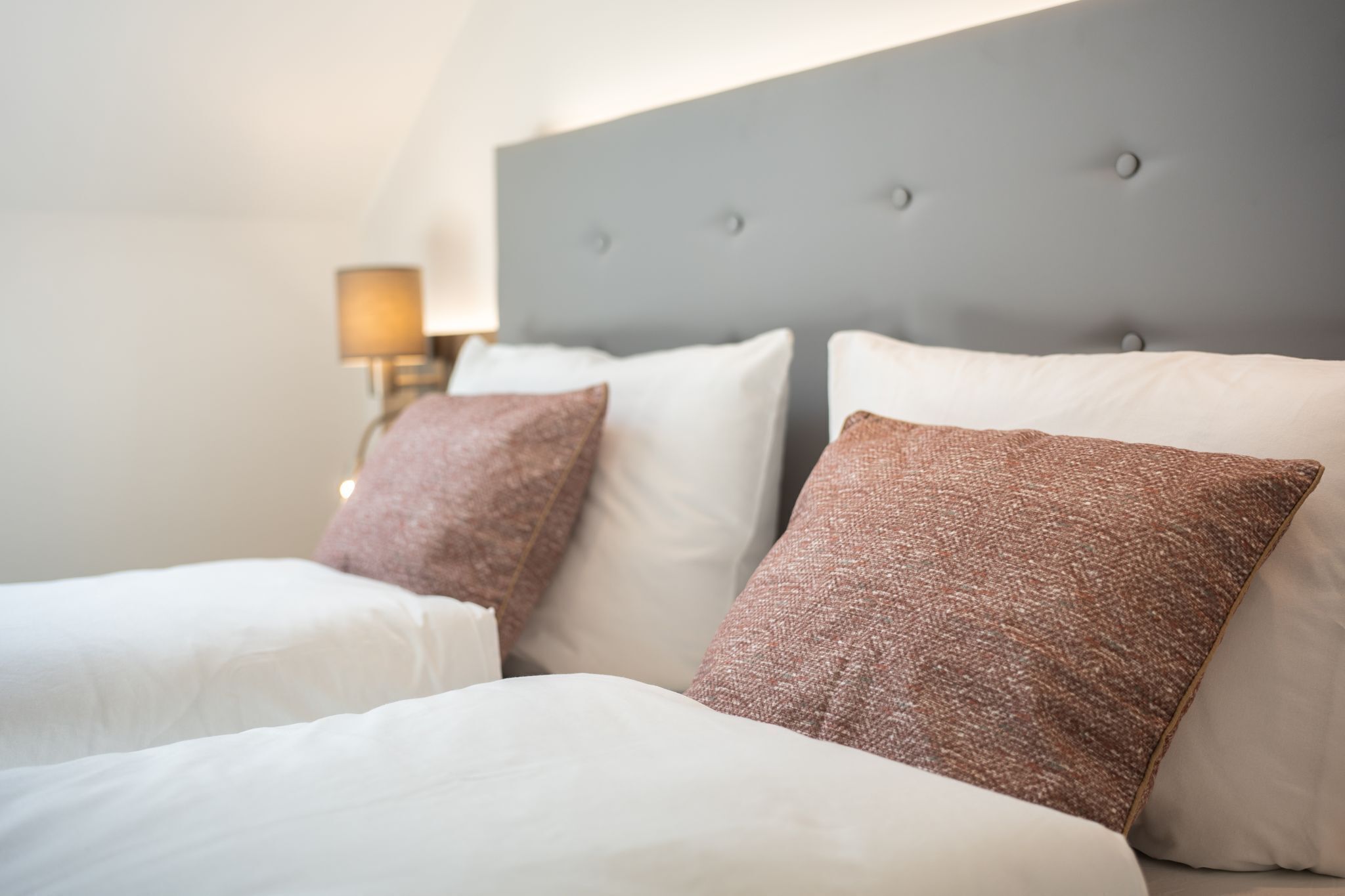 A hotel room with a grey headboard, white bedding, and two brown pillows.