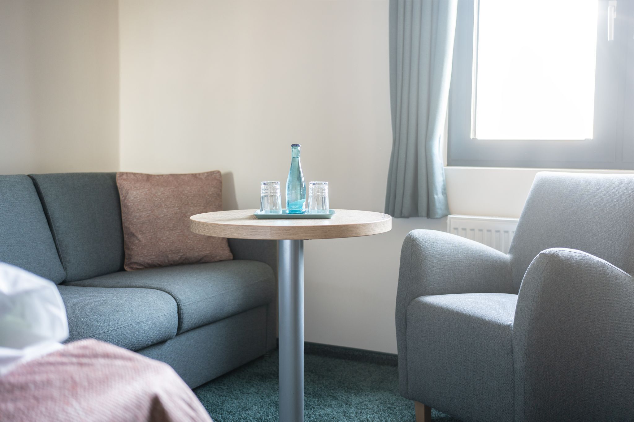 Una habitación de hotel moderna con un sofá gris, un sillón gris y una mesa redonda con una botella azul y vasos.
