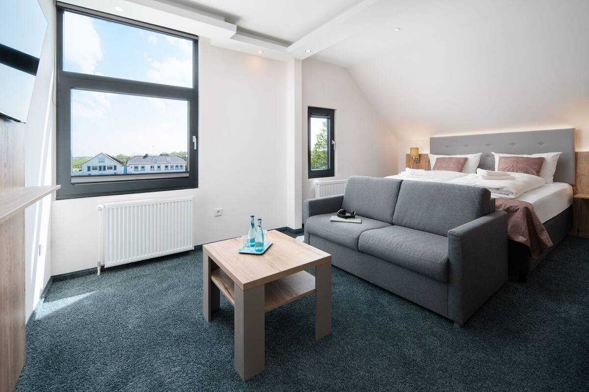 Ein modernes Hotelzimmer mit großem Fenster, einem grauen Sofa, einem Holztisch und einem Bett mit grauem Kopfteil.