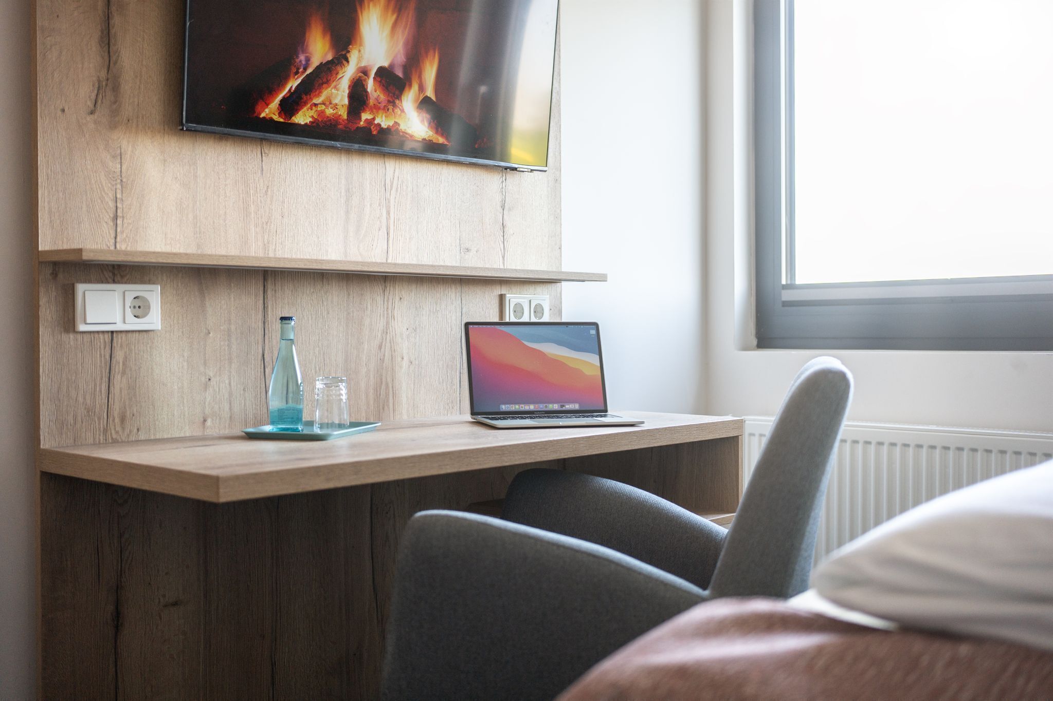 Una habitación con un escritorio de madera, una computadora portátil, una botella de vidrio y un televisor que muestra un fuego.