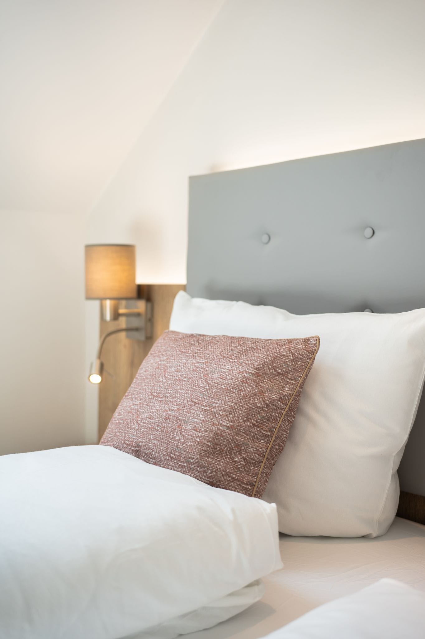 A neatly made bed with white and pink textured pillows, a grey headboard, and a bedside lamp.